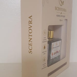 May include: A beige rectangular box for a Scentovra home diffuser containing a 100 ml glass bottle with a gold cap. The packaging features the brand name, icons for long-lasting fragrance, made with care, and elevate your atmosphere, and a label reading Warm Luxury.