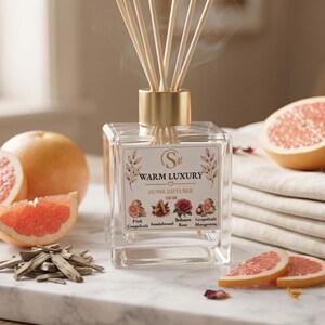 May include: A square glass reed diffuser with a gold cap and wooden reeds. The label reads WARM LUXURY HOME DIFFUSER 100 ML with illustrations of pink grapefruit, sandalwood, rose, and mangosteen. It is surrounded by grapefruit slices, wood chips, and a red rose.