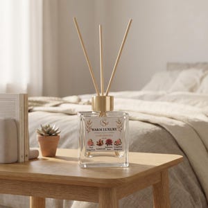 May include: A clear glass reed diffuser bottle with a gold-toned cap and three wooden reeds. The label features the text WARM LUXURY and small illustrations of flowers and fruits. The bottle is square-shaped and sits on a light wooden surface.