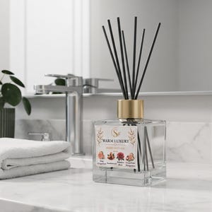 May include: A clear glass reed diffuser with a gold-toned collar and black reeds. The square bottle features a white label with the text WARM LUXURY and illustrations of flowers and fruits, including fig, sandalwood, rose, and grapefruit.