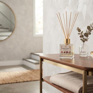 May include: A square glass reed diffuser with a gold cap and wooden sticks, a small clear glass vase with a eucalyptus sprig, and a white bowl with smooth stones, all arranged on a wooden console table with a gold metal frame and a folded blanket.