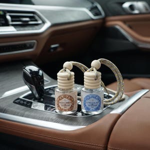 May include: Two small glass car diffuser bottles with wooden caps and hanging cords. One bottle has a brown label reading Fruity Fresh Car Diffuser 8ml, and the other has a blue label reading Citrus Blast Car Diffuser 8ml. Both feature ornate borders.