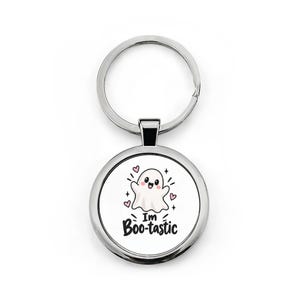 May include: A round silver metal keychain featuring a white insert with a cartoon ghost illustration, pink hearts, and the black text "I'm Boo-tastic". The design includes a standard circular split ring attached to the top of the circular metal frame.