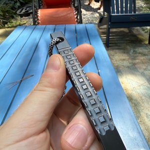 Puede incluir: Una herramienta de palanca de metal negro mate con un patrón de rejilla texturizado a lo largo del centro del eje y un orificio circular en el extremo superior. Un cordón de paracaídas con patrón blanco y negro pasa por el orificio.