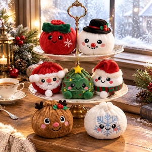 May include: A collection of seven plush holiday-themed ornaments arranged on a two-tiered white stand. The plushies feature embroidered faces and festive designs including a red ornament, a snowman, Santa Claus, a Christmas tree, and a snowflake.