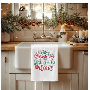 May include: A white kitchen towel featuring red and green text that reads: The joy of Christmas is family, just kidding it's wine, accompanied by a small graphic of a wine glass. The towel is draped over the edge of a white farmhouse sink.