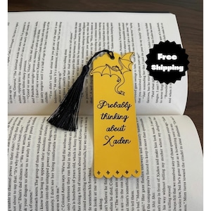 May include: A yellow rectangular bookmark with a black tassel, featuring a black line drawing of a dragon at the top and the black text "Probably thinking about Xaden" printed in the center, with scalloped edges at the top and bottom.