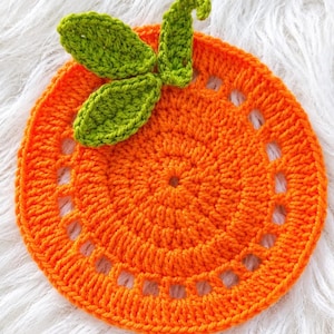 May include: A round, orange crochet coaster featuring a textured, circular pattern with a decorative openwork border. Attached to the top are three small, bright green crocheted leaves with visible stitching, creating a fruit-like appearance.