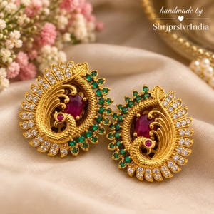 May include: A pair of gold-toned earrings featuring a peacock motif with a central oval-shaped red stone, surrounded by small green stones and clear white crystals. The metalwork has a textured finish, and the design is symmetrical.