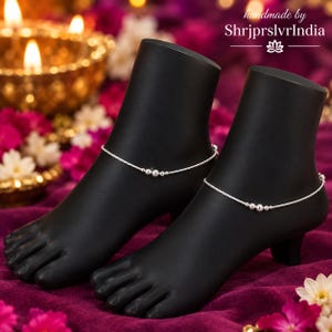 May include: A pair of silver anklets featuring a delicate chain with three small beads at the center, displayed on two black mannequin feet. The text "handmade by ShrjprslvrIndia" is visible in the upper right corner above a lotus flower icon.