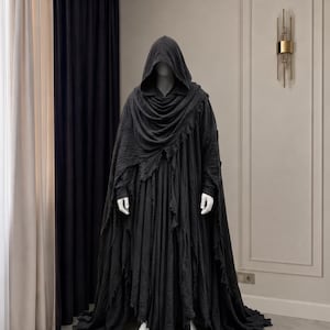 May include: A full-length, dark charcoal gray hooded cloak worn over a matching long-sleeved tunic. The garment features a layered, distressed fabric design with frayed, uneven edges along the hem and shoulders, creating a textured appearance.