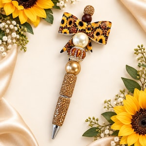 Op de afbeelding: Een decoratieve pen met een reeks gestapelde kralen in goud-, crème- en bruintinten, bekroond met een stoffen strik met een patroon van zonnebloemen en luipaardprint. De onderste houder is bedekt met kleine, sprankelende gouden kristallen en een zilveren punt.