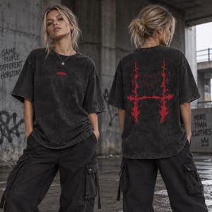 Könnte beinhalten: Ein anthrazitfarbenes T-Shirt in Acid-Wash-Optik mit einer kleinen roten Regenschirm-Grafik auf der Brust. Die Rückseite des T-Shirts zeigt ein großes, komplexes rotes Grafikdesign mit flammenartigen Mustern um ein herzförmiges Element.