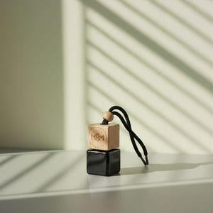 May include: A hanging car air freshener featuring a glossy black square base topped with a light-colored wooden cube. The wooden cube displays a circular logo with the letters MBM in the center. A black cord with a small wooden bead is attached.