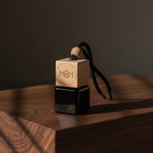 May include: A square car air freshener featuring a light wood top block engraved with the letters MGM inside a circle and a glossy black bottom block. A small wooden bead sits above the wood block, attached to a black cord for hanging.