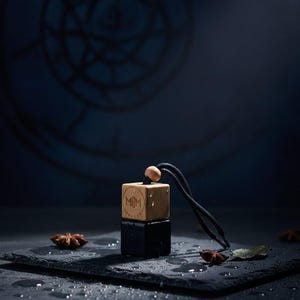 May include: A square car air freshener featuring a light-coloured wooden top block with the letters MJM engraved on the front and a solid black bottom block. A black cord with a small wooden bead is attached to the top for hanging purposes.