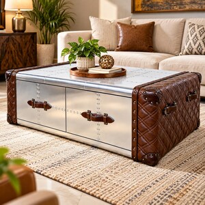 May include: A rectangular coffee table featuring a smooth silver-toned metal top and front panels with visible rivets. The sides are upholstered in brown quilted leather with metal corner reinforcements and brown leather handles on the front drawers.