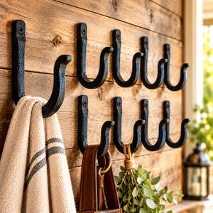 May include: A set of ten black metal wall hooks arranged in two rows, each featuring a curved J-shape and a single screw mounting point. A beige towel with dark stripes hangs from the leftmost hook, while a brown leather strap hangs from another.
