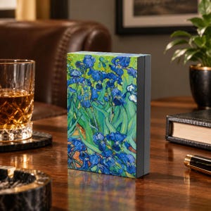 May include: A rectangular box featuring a reproduction of Vincent van Gogh's Irises painting, displaying vibrant blue flowers and green foliage on the front panel, with a solid dark gray side edge standing on a wooden surface.