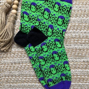 May include: A pair of knit socks featuring a vibrant neon green background with a repeating pattern of purple and black stylized figures. The socks have solid purple ribbed cuffs and solid purple toe sections, with black heel patches.