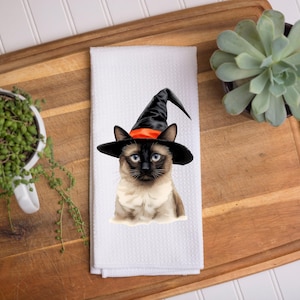 May include: A white towel features a Siamese cat wearing a black witch hat with an orange band. The cat has blue eyes and a cream-colored chest. The towel is on a wooden cutting board with a succulent and a hanging plant.