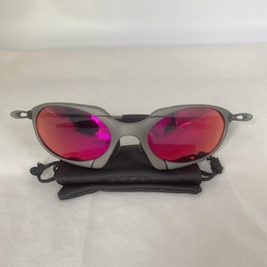 May include: Sunglasses with a matte grey metal frame and reflective pink lenses. The sunglasses are positioned directly on top of a small, black, rectangular drawstring pouch made of fabric, featuring a subtle embossed logo on the front surface.