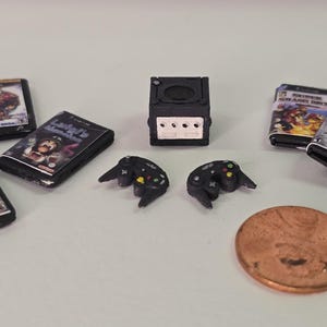 May include: A miniature black game console, two black game controllers, and five small game cases with printed covers, including titles like Luigi's Mansion and Crash Bandicoot, arranged next to a copper-colored United States penny for scale.