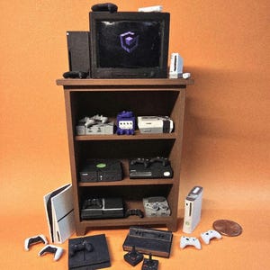 May include: A miniature brown wooden shelving unit displaying various small-scale video game consoles and controllers. A television sits on top showing a purple logo. A copper penny is placed nearby for scale, showing the tiny size of the items.
