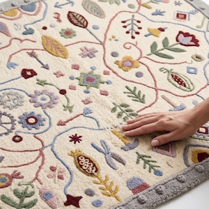 Puede incluir: Una alfombra de color crema con un patrón bordado multicolor de flores, hojas y formas geométricas. La alfombra tiene un pelo grueso y texturizado, y un borde ancho de color gris con un diseño de puntos circulares repetidos.