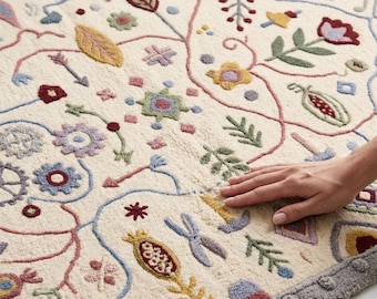 Könnte beinhalten: Ein cremefarbener getufteter Teppich mit einem mehrfarbigen Stickmuster aus Blumen, Blättern und geometrischen Formen. Der Teppich hat einen dicken, strukturierten Flor und einen breiten, grauen Rand mit einem wiederkehrenden Punktmuster.