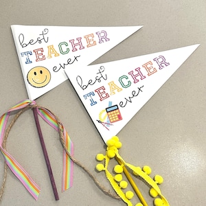 Puede incluir: Dos banderines blancos con el texto "mejor profesor de todos los tiempos" en letras de colores. Una bandera tiene una cara sonriente y la otra una calculadora y otros útiles escolares. Las banderas están unidas a palos de madera con una cinta de colores y un ribete de pompones amarillos.