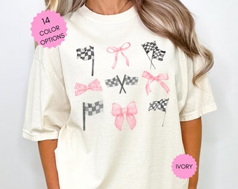 Puede incluir: Una camiseta de color crema con un gráfico de lazos rosas y banderas a cuadros en blanco y negro. El texto "14 COLOR OPTIONS" está en un círculo rosa y el texto "IVORY" está en un círculo rosa.