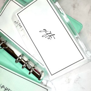 Puede incluir: Un archivador verde menta con un bolsillo de pl&aacute;stico transparente que contiene una tarjeta blanca con la palabra "regalos" escrita en cursiva negra. El archivador tiene un mecanismo de anillas plateado y una tarjeta blanca con la palabra "mejora" escrita en cursiva negra es visible debajo del bolsillo.