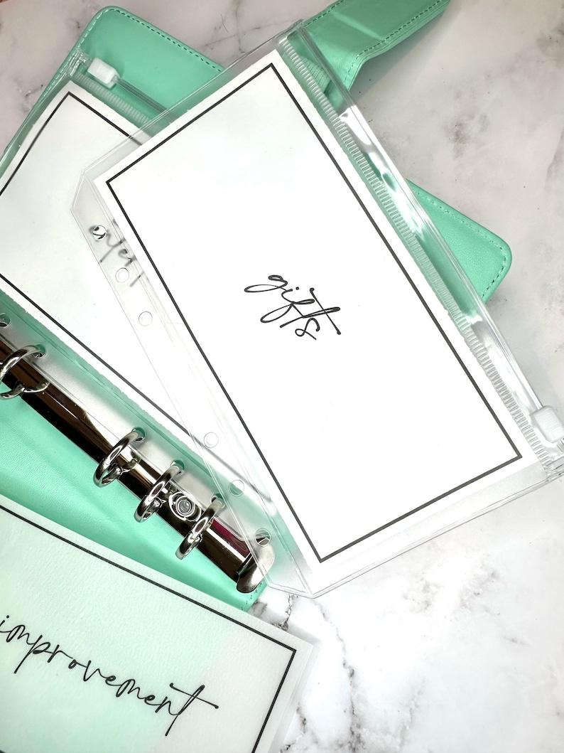 Puede incluir: Un archivador verde menta con un bolsillo de pl&aacute;stico transparente que contiene una tarjeta blanca con la palabra "regalos" escrita en cursiva negra. El archivador tiene un mecanismo de anillas plateado y una tarjeta blanca con la palabra "mejora" escrita en cursiva negra es visible debajo del bolsillo.