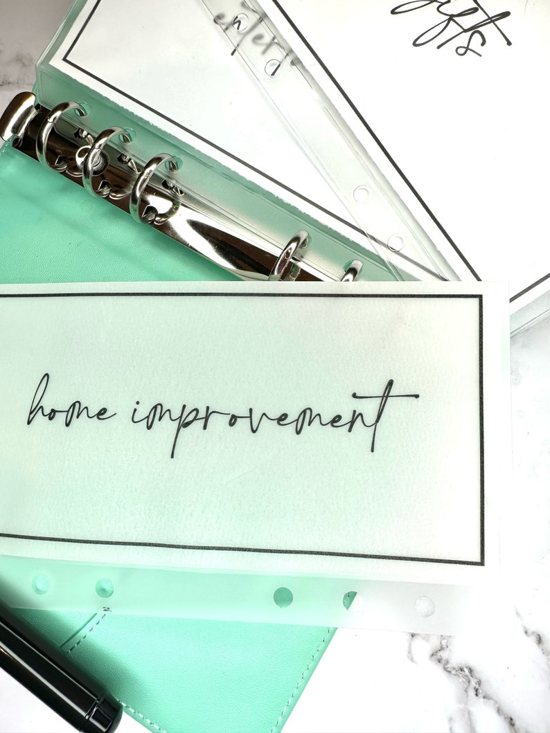 Puede incluir: Una agenda de cuero verde claro con un encuadernador de anillos de plata y un protector de p&aacute;gina de pl&aacute;stico transparente con el texto "home improvement" escrito en negro.