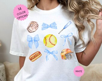 May include: White t-shirt with a softball theme. The shirt features a softball, bat, glove, hot dog, and bows in blue and white.