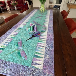 May include: A teal and purple table runner with a dragonfly design. The runner is on a wooden table with a vase of flowers and sewing thread.