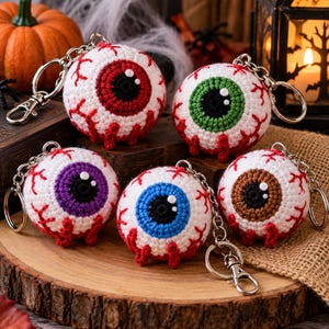 May include: Five spherical crochet keychains shaped like eyeballs, each featuring a white base with red stitched veins and a central iris in red, green, purple, blue, or brown. Each keychain includes a silver metal ring and a lobster clasp.