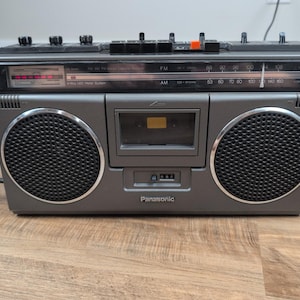 Puede incluir: Una radio y reproductor de casetes portátil Panasonic de color gris carbón con dos altavoces circulares, una platina de casete central y un panel de control superior. La pantalla frontal muestra escalas de frecuencia AM y FM.