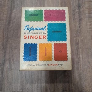 Op de afbeelding: Een rechthoekige doos voor een professionele knoopsgatmaker van Singer. De witte doos bevat kleurrijke stoffen stalen in groen, roze, blauw, oranje en rood, elk met een gestikt knoopsgat. De tekst luidt: Professional Buttonholer by Singer. What's new for tomorrow is at SINGER today!