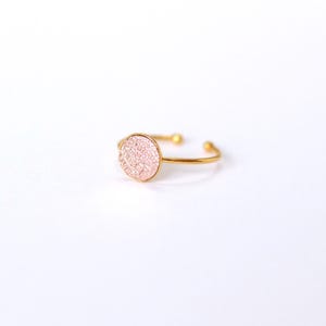 May include: A gold-toned open ring featuring a circular centrepiece with a textured, shimmering light pink surface. The band is thin and metallic, ending in two small spherical terminals at the opening, creating a delicate and adjustable design.