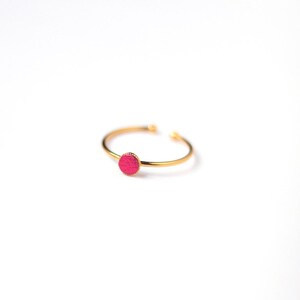 May include: A fine gold-coloured ring with a small, circular, bright pink feature. The ring has an open design, allowing for slight size adjustment. The vivid pink colour of the accent provides a striking contrast against the gold band. This ring is a simple yet elegant piece of jewellery.
