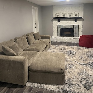 May include: A large, light brown sectional sofa with a matching ottoman, positioned on a textured gray and white area rug. The living room features a white stone fireplace with a black mantel and a red beanbag chair in the corner.