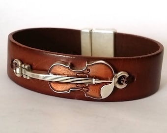 May include: A brown leather bracelet with a silver magnetic clasp and a silver violin charm. The violin charm is detailed with a brown wood grain pattern.