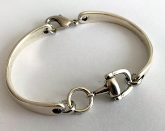 May include: Silver bracelet with a horse bit design. The bracelet has a curved, smooth band and a clasp. The horse bit detail is central, adding an equestrian touch to the jewellery. The bracelet is approximately 18 cm in length.