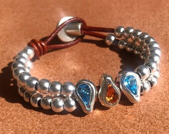 May include: A silver beaded bracelet with a brown leather strap and three teardrop-shaped charms with blue and orange gemstones.