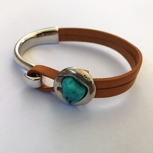 Può includere: Un braccialetto in pelle marrone con chiusura in argento e un accento in turchese.