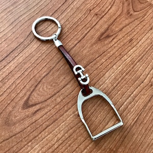 May include: A silver stirrup-shaped keyring with a brown leather strap and a silver ring. The stirrup has a small silver detail in the shape of a horseshoe.