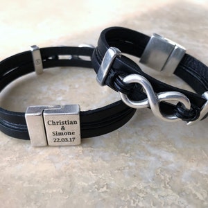 May include: Two black leather bracelets with silver infinity symbol charms and a silver clasp. One bracelet has the text "Christian & Simone 22.03.17" engraved on the clasp.