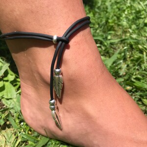 Op de afbeelding: Een zwarte enkelband met twee zilveren verencharms. De armband is gemaakt van een rekbaar koord en is verstelbaar.